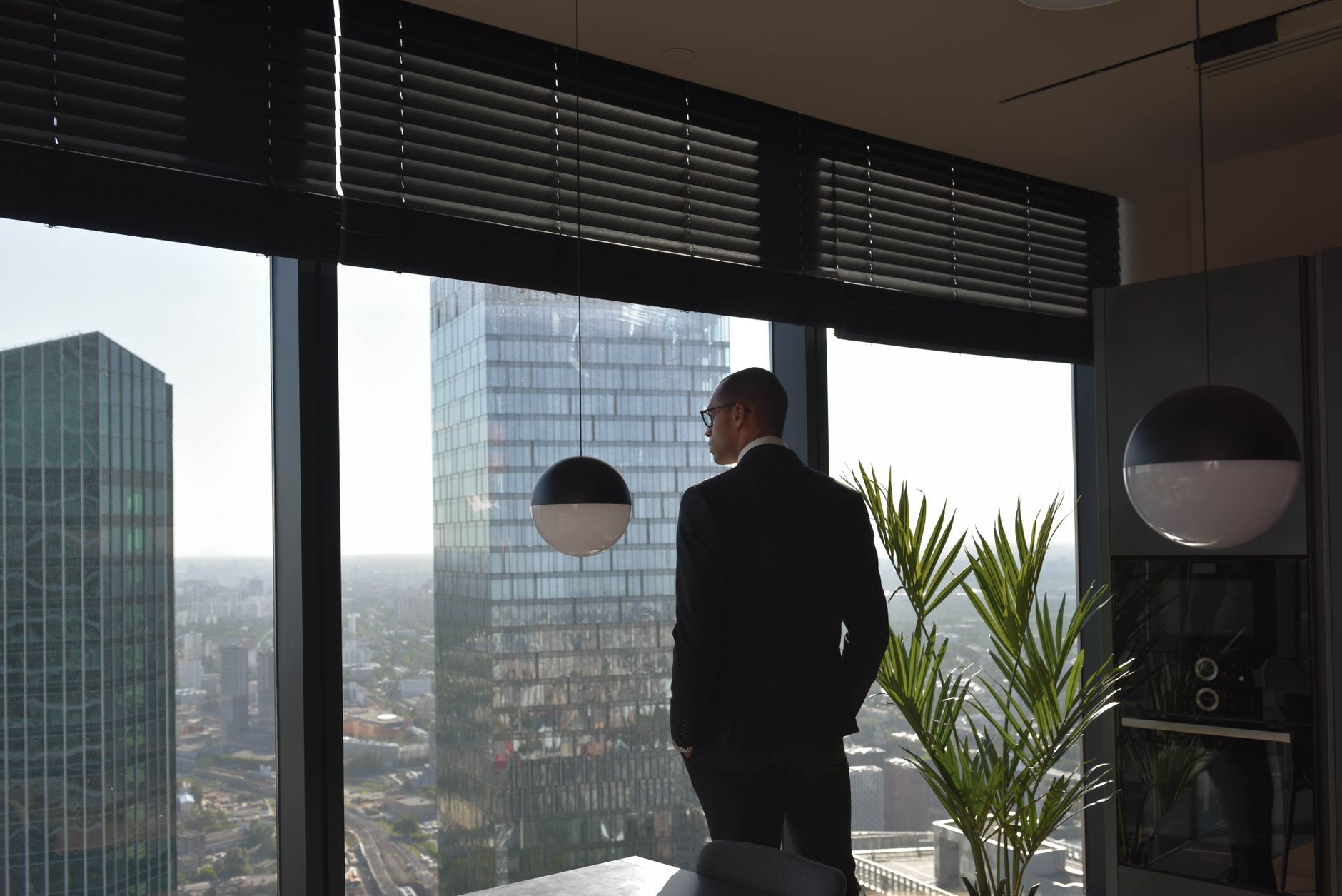 man in black suit standing beside window and looking at skyscrapers